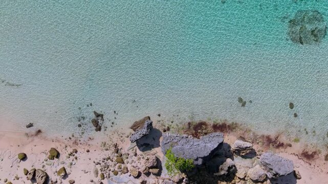 Top down aerial view of crystal clear turquoise sea water gently lapping a sandy beach with rocks and coral reefs