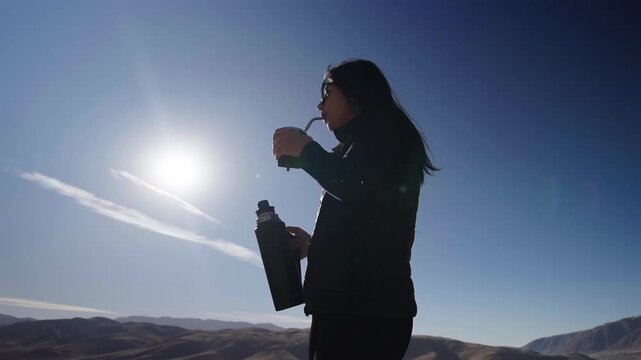 Woman drinking yerba mate in Taf&iacute; del Valle, Tucum&aacute;n, Argentina, silhouetted against mountain sunrise with traditional stainless steel thermos and Andean landscape backdrop, slow motion shot