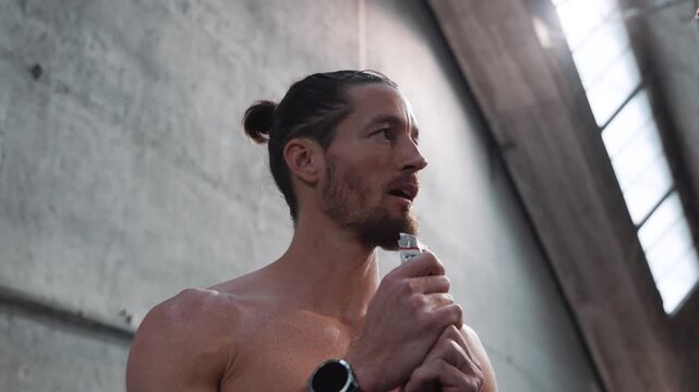 A low-angle shot of a muscular, shirtless young man with his hair tied back and a beard, wearing a watch on his right wrist, eating a sports energy gel