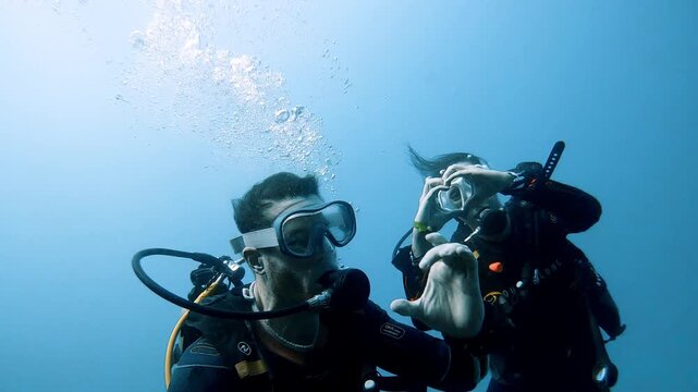 A male and female diving duo underwater in full gear; the woman is forming a heart with her hands and the man a half-heart