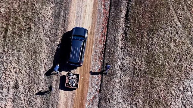 Aerial view of a black SUV towing a small trailer on a rural dirt road while workers stand beside a gravel work area.