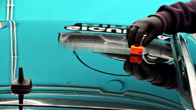 Gloved hand applying detailing tool on a glossy turquoise car hood, showing paint protection work and reflective auto studio finish.