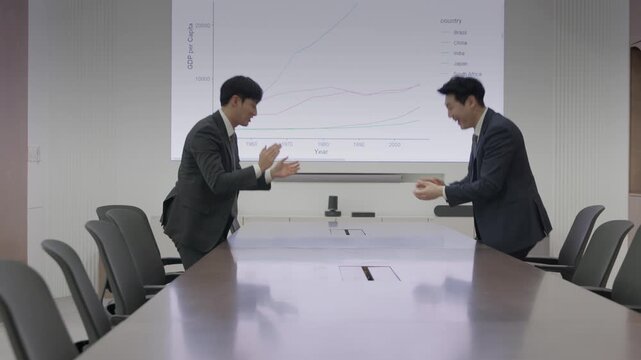 Two excited businessmen in suits celebrating a business success with raised fists and big smiles in front of a projection screen displaying a GDP line chart in a modern boardroom.