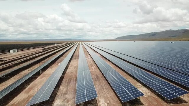 An expansive field of solar panels on a sunny day, viewed from a side angle. The solar panels have a bluish, glossy tint, positioned side by side in a sandy field on a cloudy yet sunny day.