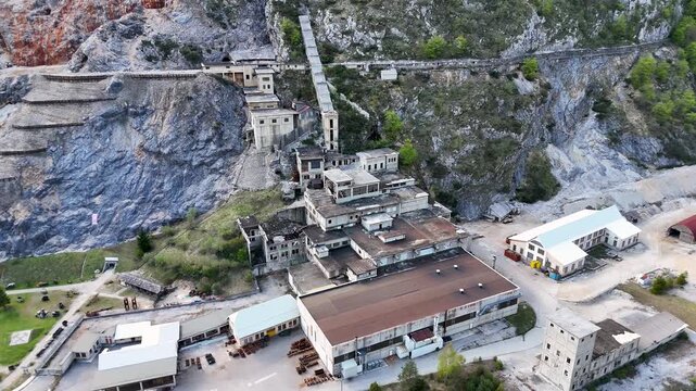 Drone view of a quarry wall, industrial buildings, and Tarvisio village framed by steep forested alpine mountains in northern Italy.