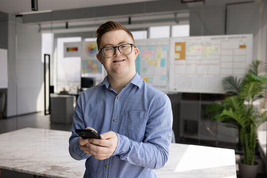 Portrait of young 25s smiling man with Down syndrome standing alone in modern office, using smartphone. Inclusion, accessibility, digital technology and equal opportunities for people with disability