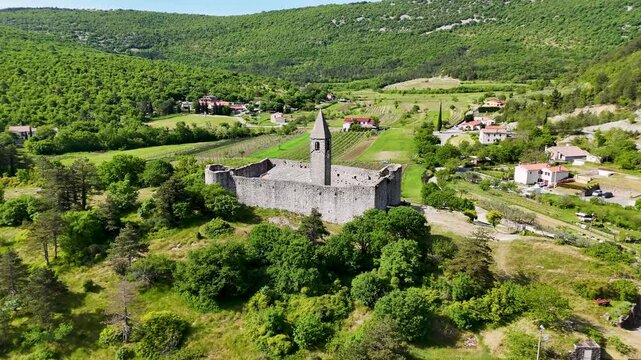 Close drone view of Cerkev sv. Trojice in Hrastovlje, Slovenia, showing stone walls, bell tower, slate roof, and surrounding greenery.