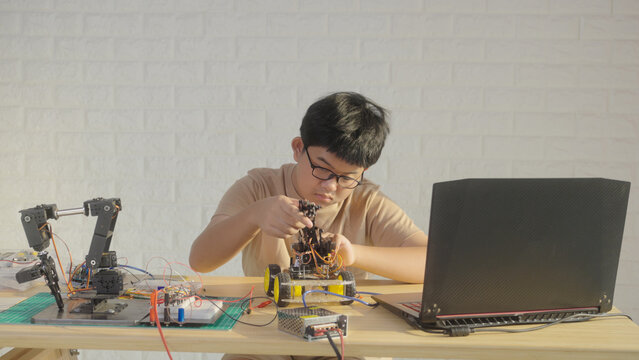 Asian young student building a robotics project at home with a robot arm, electronics, and laptop. Concept of STEM education, engineering skills, innovation, and future technology learning.