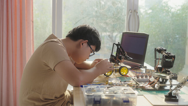 Asian young student assembling a robotics vehicle at home with electronics, robotic arm, and laptop. Concept of STEM education, engineering skills, innovation, and hands-on technology learning.
