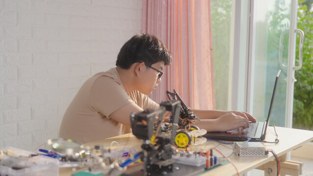 Asian young student using a laptop at home while working on a robotics project with electronic components and robotic devices. Concept of STEM education, coding, and technology learning.