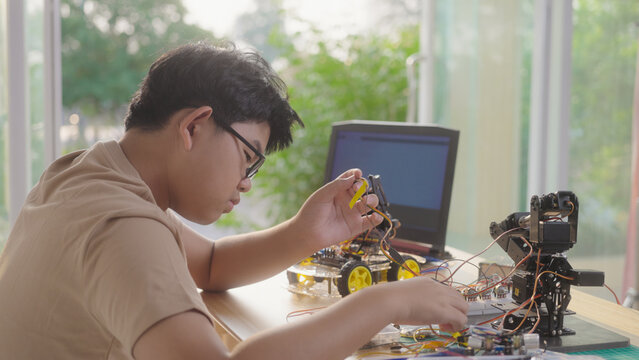 Asian young student wiring a robotics project at home with robotic devices, electronics, and a laptop. Concept of STEM education, engineering skills, innovation, and hands-on technology learning.