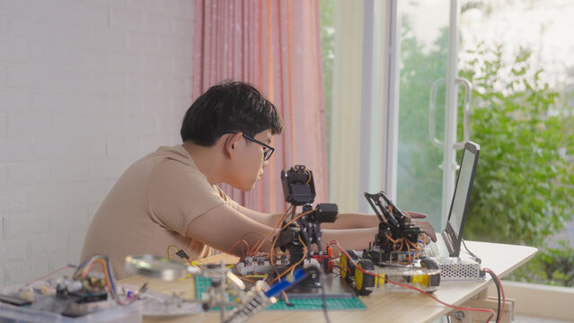 Asian young student using a laptop at home while working on a robotics project with electronic components and robotic devices. Concept of STEM education, coding, and technology learning.