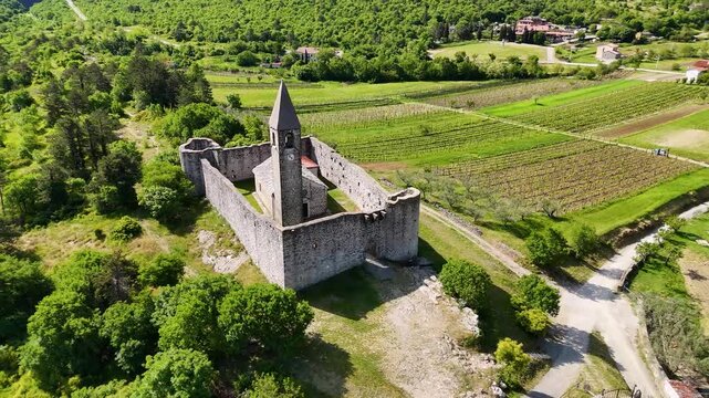 Top-down drone view of the fortified Holy Trinity Church in Hrastovlje, Slovenia, showing stone walls, bell tower, courtyard, and vineyards.