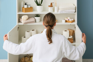 Naklejka premium Woman in white robe organizing bathroom cabinet with toilet paper and personal care products, home organization and storage