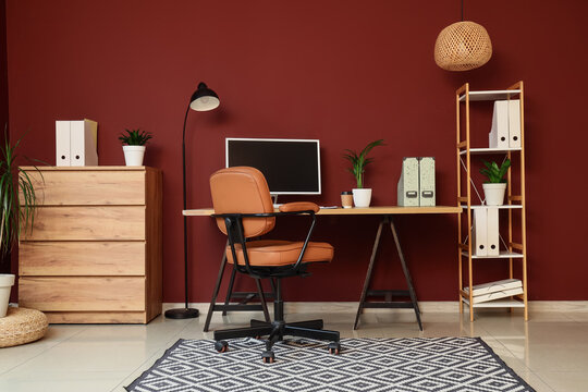 Modern home office interior with computer monitor, brown leather chair, wooden desk, and red wall decor