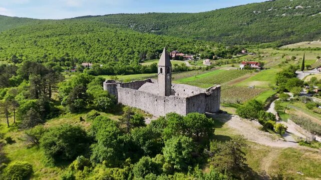 Drone view of the fortified Holy Trinity Church in Hrastovlje, Slovenia, surrounded by fields, homes, trees, and rolling Istrian hills.