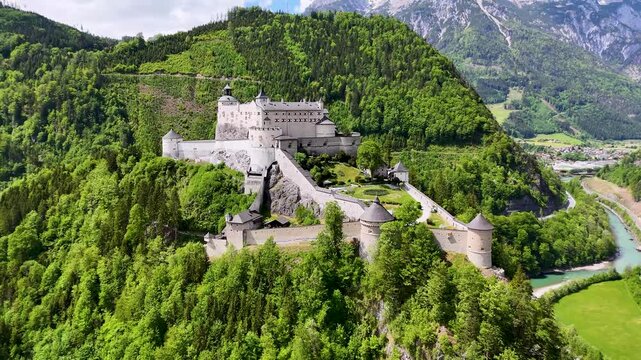 Drone side view of Burg Hohenwerfen fortress on a wooded cliff in Werfen, Salzburg, Austria, with mountains and valley beyond.