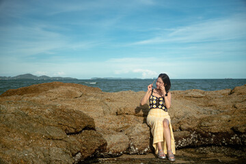 Fototapeta premium A young Asian woman wearing a floral crochet crop top and sunglasses sitting and posing on a rock formation at a beach with the ocean background