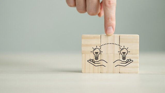A hand places a wooden block with two lightbulb designs on a table indoors.