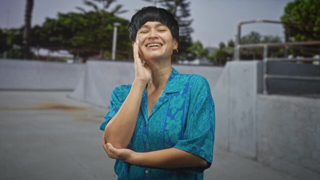 Woman smiling with hand on cheek, head tilted and wearing blue floral button shirt, hoop earring and short haircut on a concrete street beside a skatepark wall and metal railing; contentment.