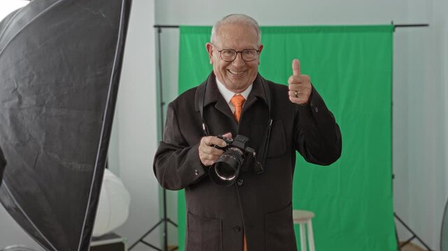 Senior man photographer holding a dslr camera and giving thumbs up in a studio with green screen and softbox lighting; confidence cheerful.