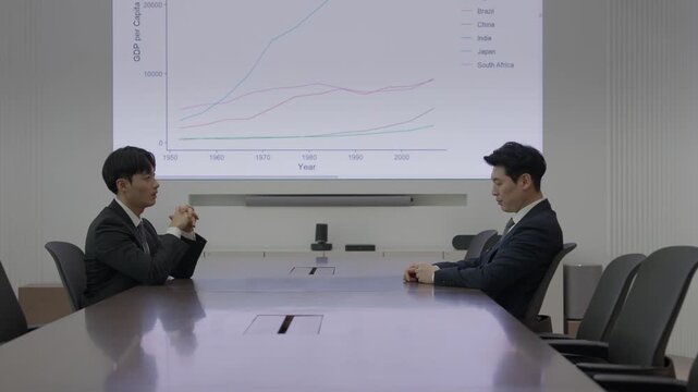 Two businessmen in dark suits sitting across from each other at a large conference table reviewing a projected GDP line chart during a formal business meeting in a modern boardroom.