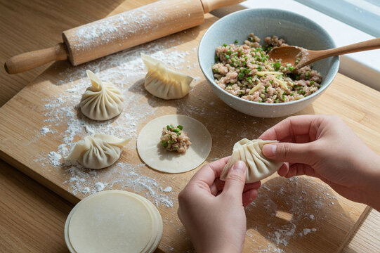 Hands folding and pleating fresh dumpling wrappers on a wooden board with bowl of filling and rolling pin