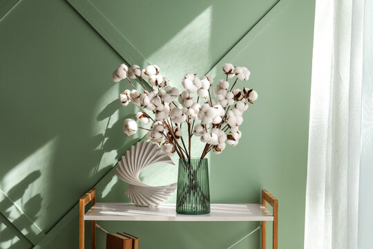 Fluffy white cotton bolls in green ribbed vase on white shelf with books and modern decor, the shelf also holds two brown books and a white modern decorative object