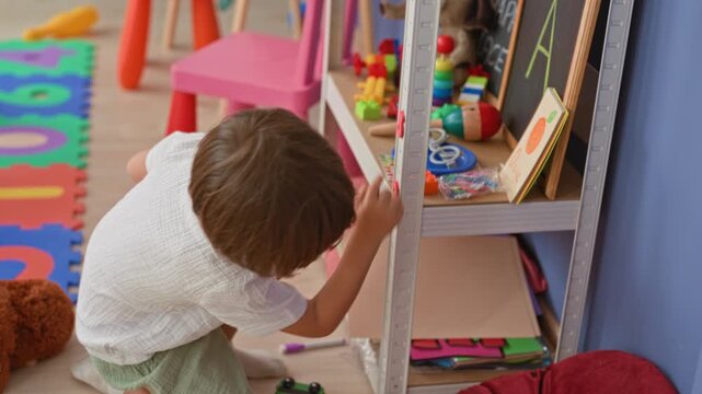 Boy kneeling and reaching, finger touching a toy on a low shelf beside colorful foam mat and stuffed bear inside a building; curiosity.