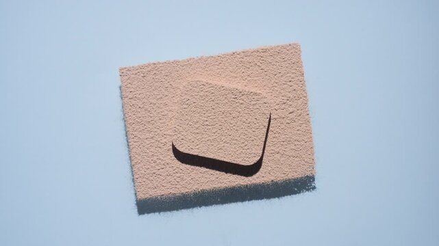 Top view shot of beige fine powder sifted into a large rectangular shape with a smaller rectangular pedestal at the center, casting deep, dynamic shadows under orbiting light on a blue background | 