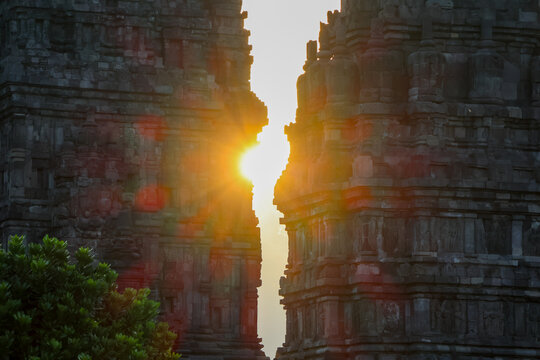 Sunlight pierces ancient stone towers Prambanan temple. Warm glow bathes weathered, towering ruins. Rugged carvings stand silent through time. Nature's green whispers at base of stone