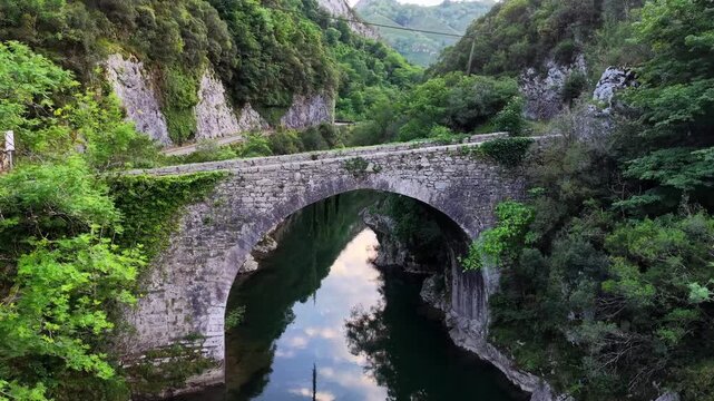 Old stone arch bridge in rural area of Picos de Europa mountains national park area. Drone shot slowly rising up for high angle view of stone arch bridge over river in northern Spain.