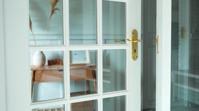 A modern white door with gold handle reflects a wooden table and vase through its glass panels indoors.