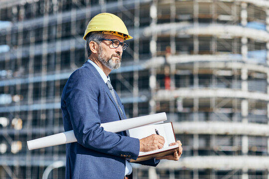 senior businessman architect wearing a protective helmet writing in a notebook and holding a blueprint on the construction site