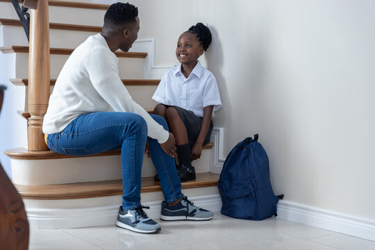 African American dad helping school-age daughter in uniform on stairs tying shoe near navy backpack