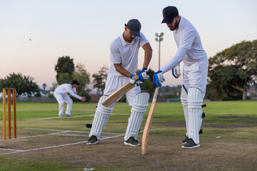 Obraz premium Two male cricketers in white kit leaning while adjusting bat grip near stumps at dusk