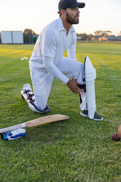 Male cricketer kneeling, adjusting leg pad on grassy outfield in white kit with bat, gloves, ball