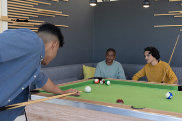 Obraz premium Three male friends playing pool on green felt table in lounge corner, watching balls, holding cues
