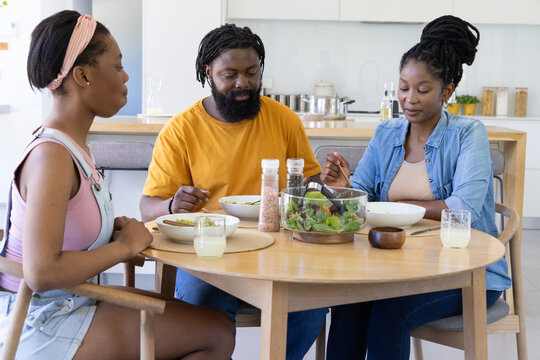 African family serving salad using glass bowl and tongs at round wooden table in kitchen