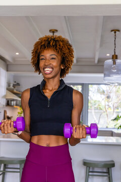 African American woman exercising with purple weights in kitchen in black top and purple leggings