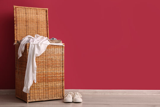 Wicker laundry hamper with dirty clothes and white sneakers on wooden floor against red wall, the background features a solid red wall, providing ample copy space