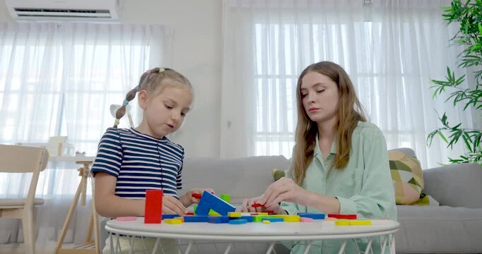 Caucasian single mother sits near preschool daughter at round table, guiding colorful block building, math practice, patient home education, nurturing care, creative childhood development indoors.