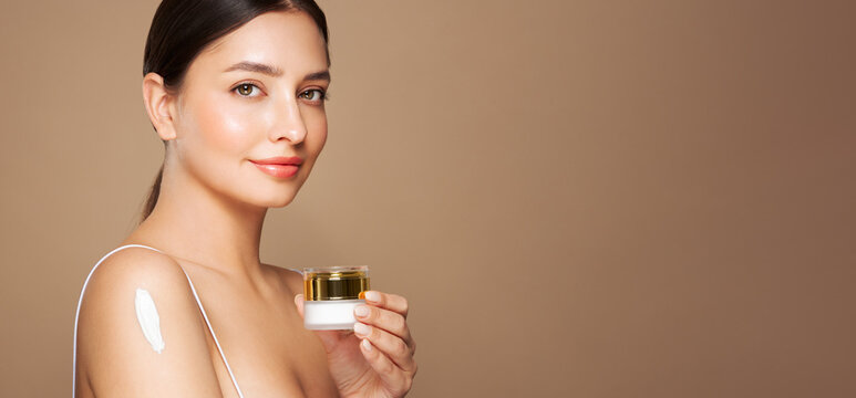Cropped shot of a young tanned woman moisturizing her shoulder with her hand using a cream isolated on a beige background. Skin care. A white thick spear of cream on the female body
