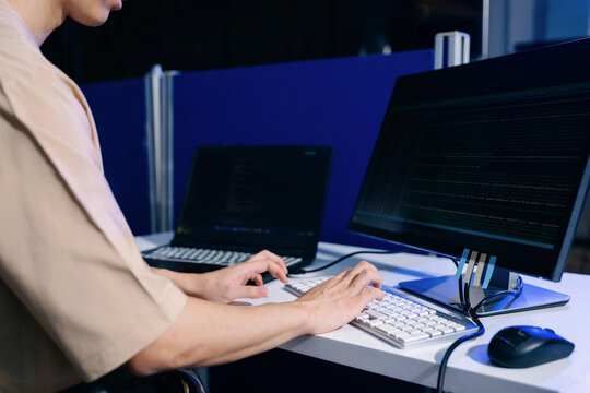 Closeup of a software developer typing data lines on a white keyboard with a monitor showing code setup.