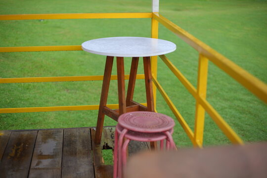 Cozy outdoor cafe table and stool on a wooden terrace overlooking a rainy, lush green grass field.