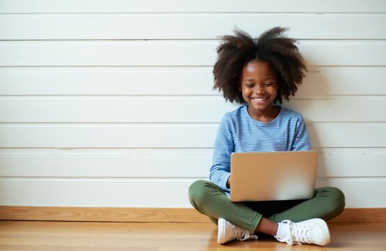Smiling young black girl learns online using a laptop. Child sits cross-legged on floor typing keyboard, bright smile, focused on education. Technology use for kids learning.