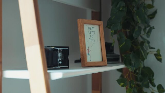 A framed quote sits on a white shelf beside a black box and a plant with green leaves.