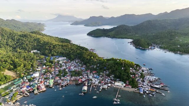 Drone footage with horizontal panning captures Liminangcong village stretched along the coast, showcasing stilt houses and hillside homes with Mountain Capoas