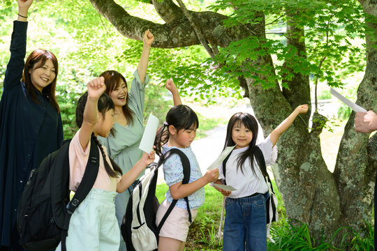 新緑の木の下で先生と一緒に右手を挙げて気合いを入れる親子と女子小学生たちのグループ