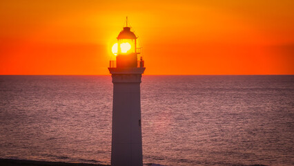 AERIAL, LENS FLARE, CLOSE UP: Lighthouse tower with the setting sun shining behind the lantern room...